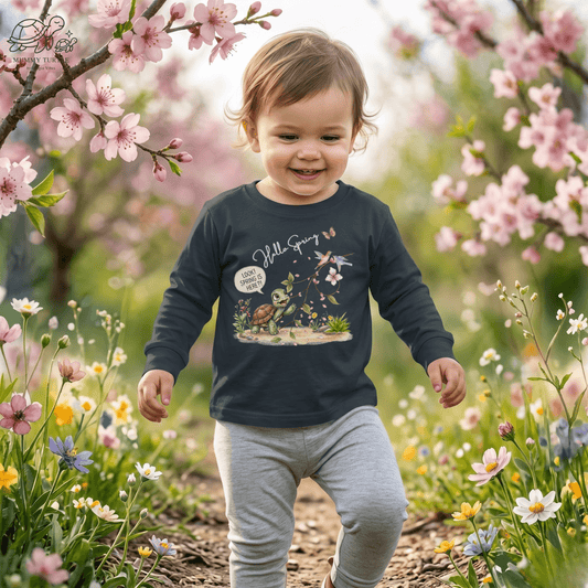 Adorable toddler in a Hello Spring toddler spring long sleeve shirt playing among blossoms in a vibrant spring garden.